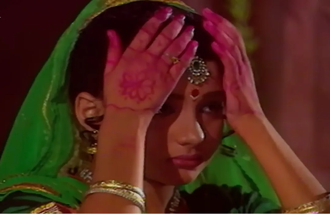 Radha Rani, draped in a vibrant green veil and adorned with gold bangles, gently raises her henna-stained hands to her forehead. With eyes c…